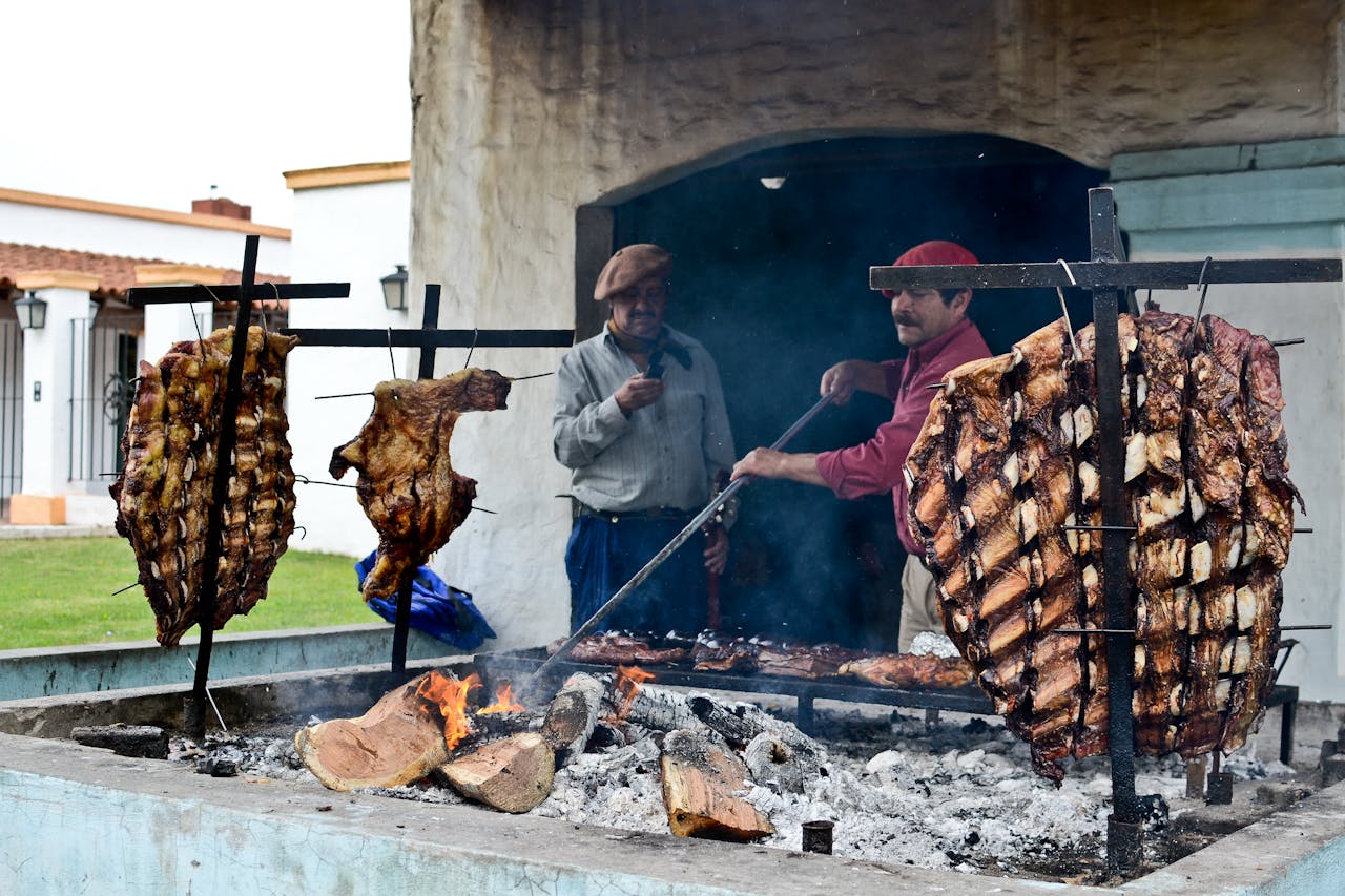 Argentine men preparing traditional asado with meats grilling over open flame outdoors.